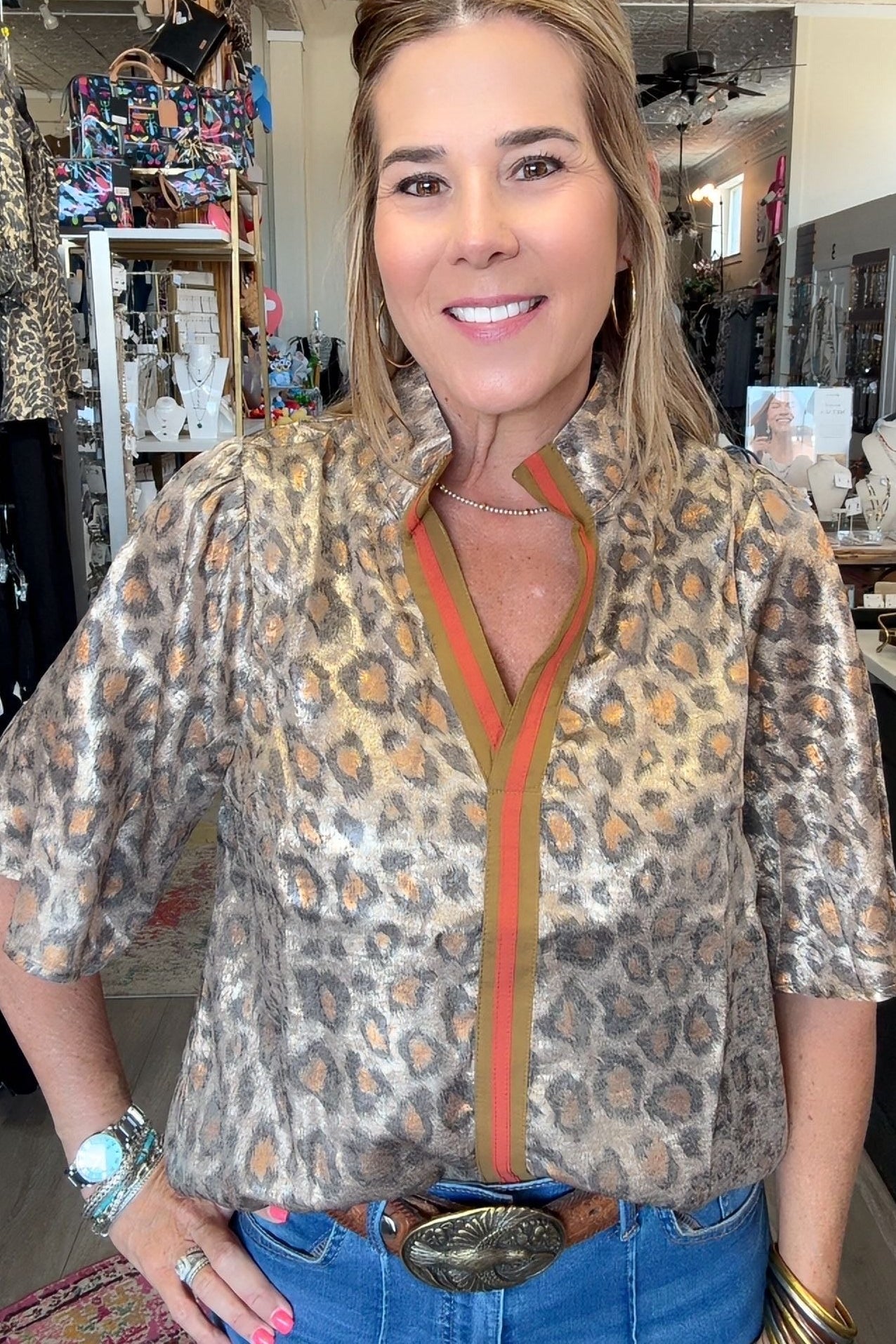A woman modeling a leopard-print blouse with a green and orange striped collar placket, paired with blue jeans and a large silver belt buckle, standing in a retail store.