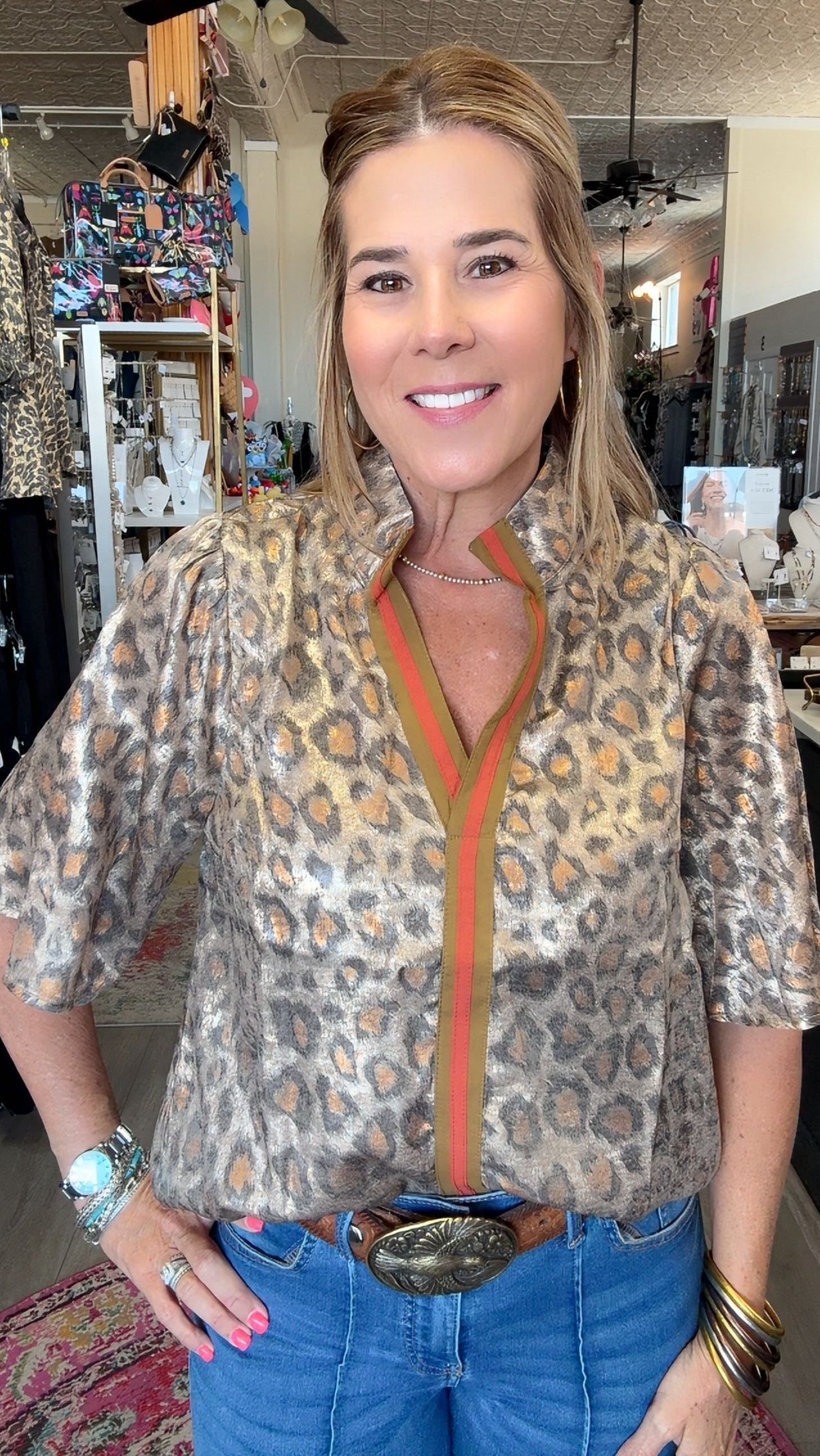A woman modeling a leopard-print blouse with a green and orange striped collar placket, paired with blue jeans and a large silver belt buckle, standing in a retail store.
