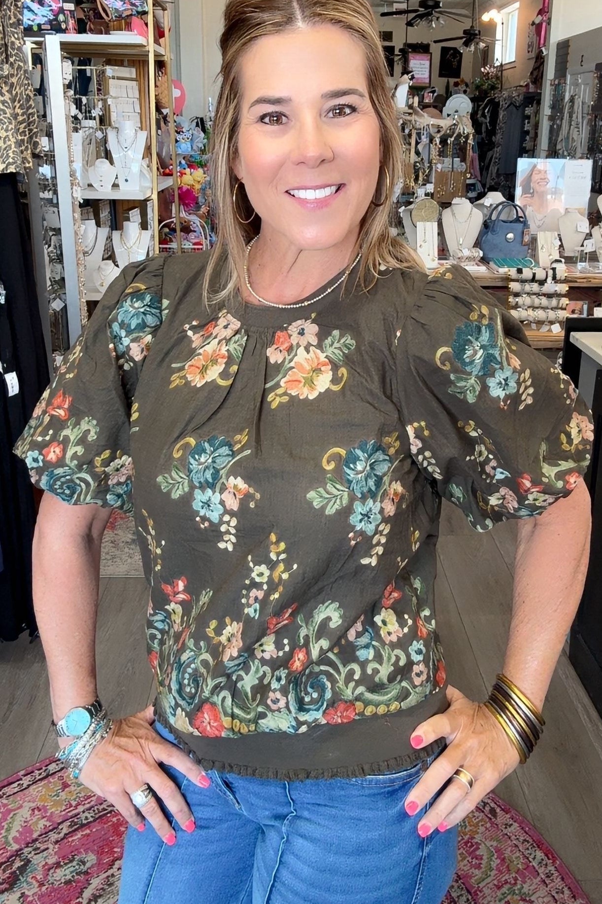 A woman posing in a boutique shop wearing a dark olive green puff-sleeve blouse with a colorful floral pattern and blue jeans.