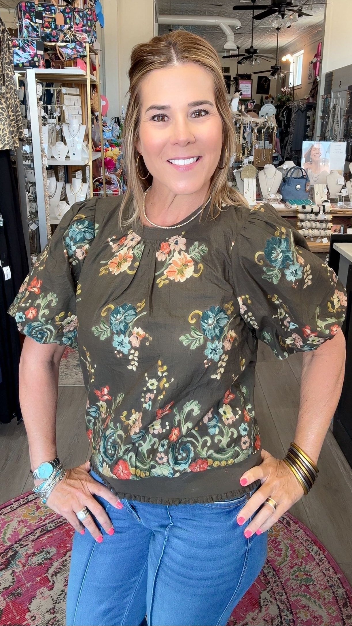 A woman posing in a boutique shop wearing a dark olive green puff-sleeve blouse with a colorful floral pattern and blue jeans.