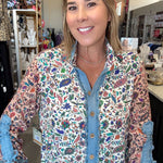 A woman wearing a long-sleeve button-up shirt with a colorful floral and bird print over a blue denim shirt, standing in a retail store.