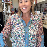 A woman wearing a long-sleeve button-up shirt with a colorful floral and bird print over a blue denim shirt, standing in a retail store.