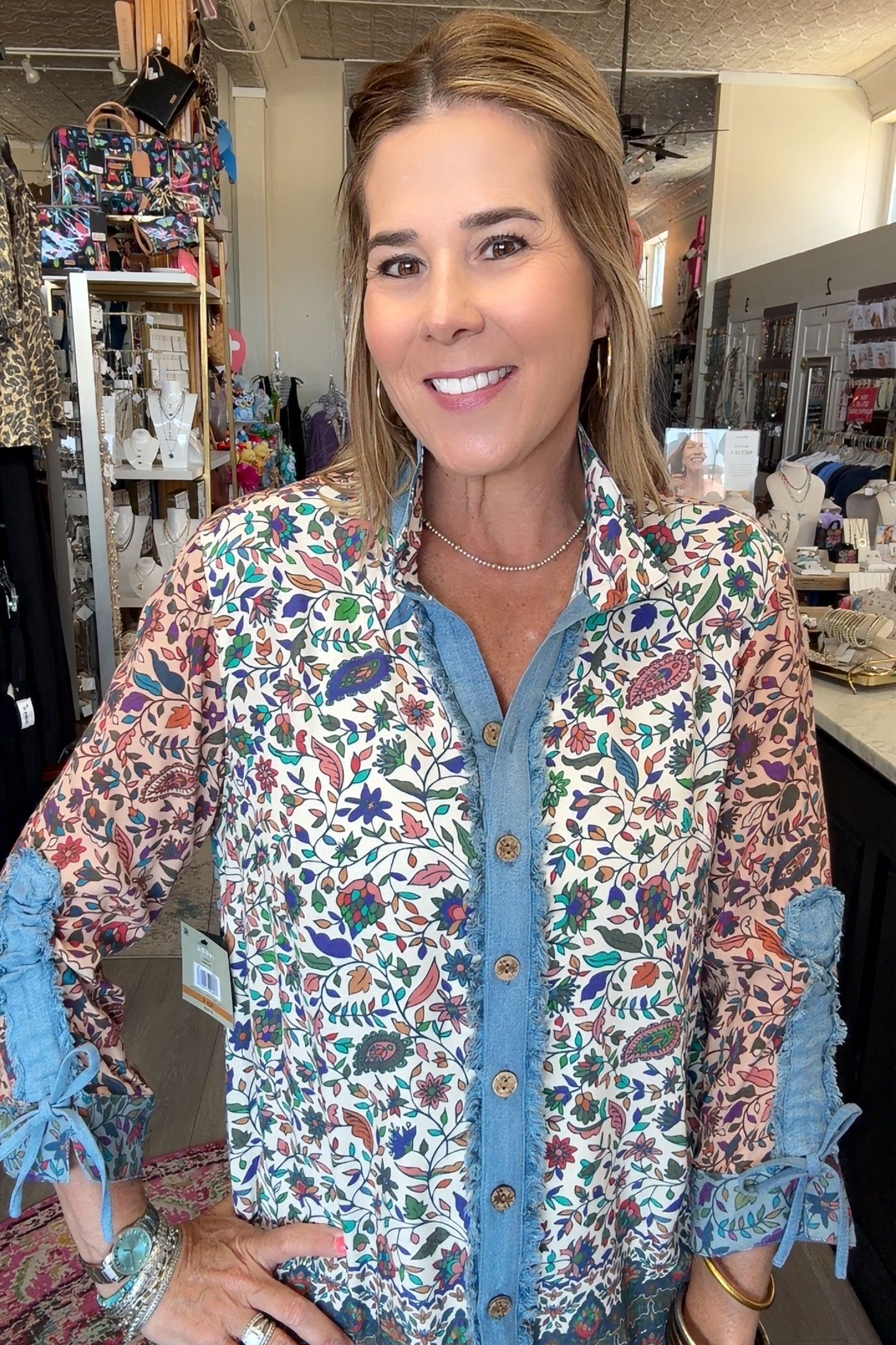 A woman wearing a long-sleeve button-up shirt with a colorful floral and bird print over a blue denim shirt, standing in a retail store.