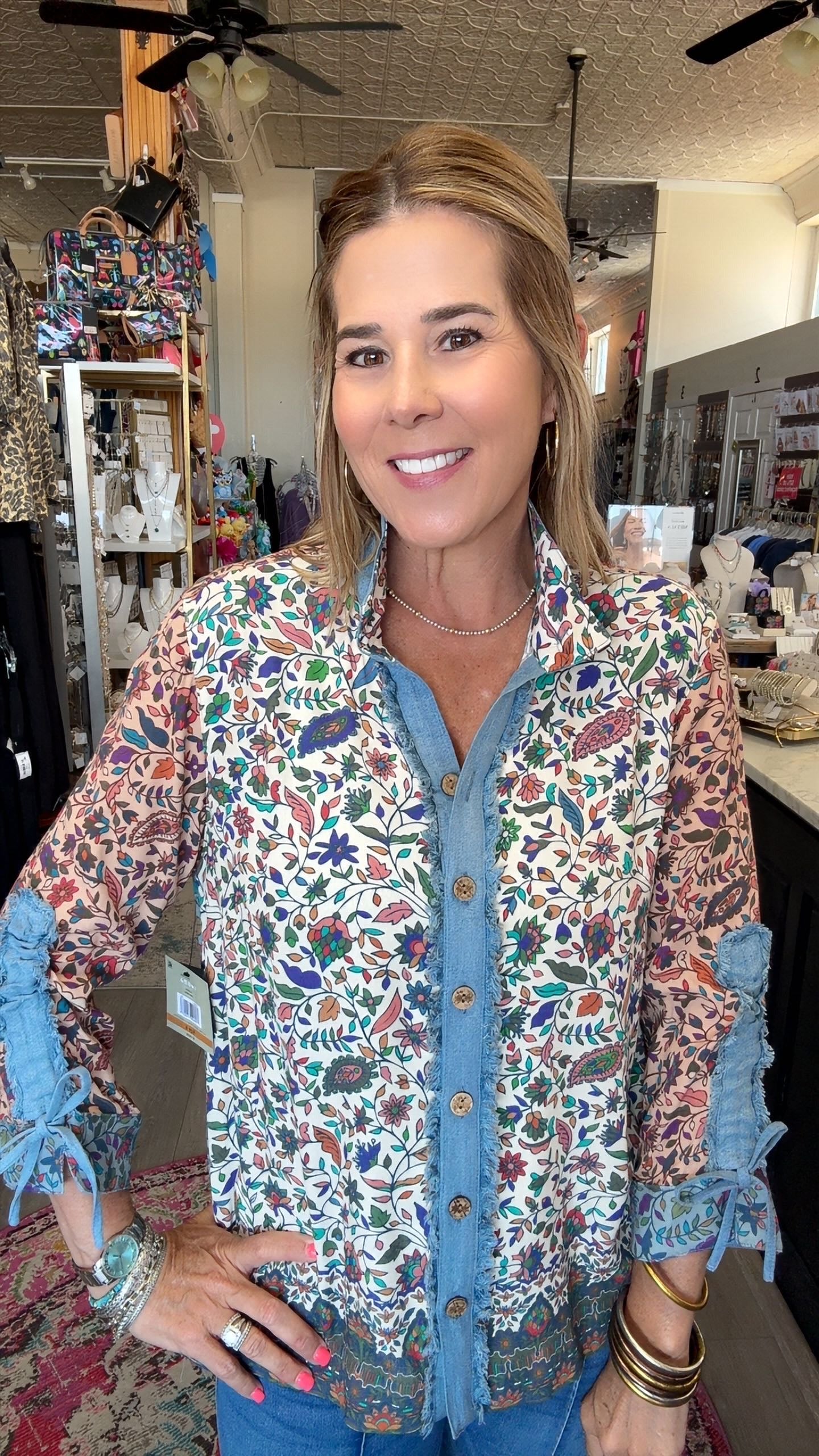 A woman wearing a long-sleeve button-up shirt with a colorful floral and bird print over a blue denim shirt, standing in a retail store.