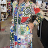 A woman modeling a long-sleeve patchwork maxi dress inside a boutique shop. The dress features a vibrant multicolor patchwork pattern with geometric and floral motifs in green, blue, red, yellow, and white.