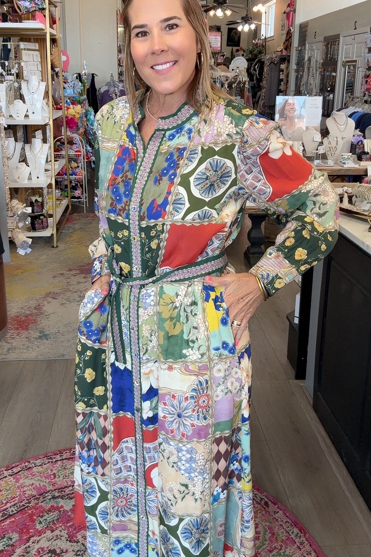 A woman modeling a long-sleeve patchwork maxi dress inside a boutique shop. The dress features a vibrant multicolor patchwork pattern with geometric and floral motifs in green, blue, red, yellow, and white.