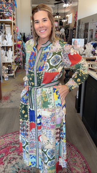 A woman modeling a long-sleeve patchwork maxi dress inside a boutique shop. The dress features a vibrant multicolor patchwork pattern with geometric and floral motifs in green, blue, red, yellow, and white.