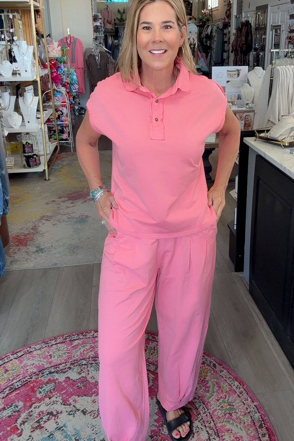 A woman modeling a matching pink two-piece lounge set consisting of a short-sleeve polo shirt and wide-leg pants, standing inside a retail boutique.