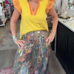 A woman posing inside a boutique shop, wearing a bright yellow ruffle-sleeve top with orange trim and wide-leg, multi-colored abstract print pants.