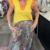 A woman posing inside a boutique shop, wearing a bright yellow ruffle-sleeve top with orange trim and wide-leg, multi-colored abstract print pants.