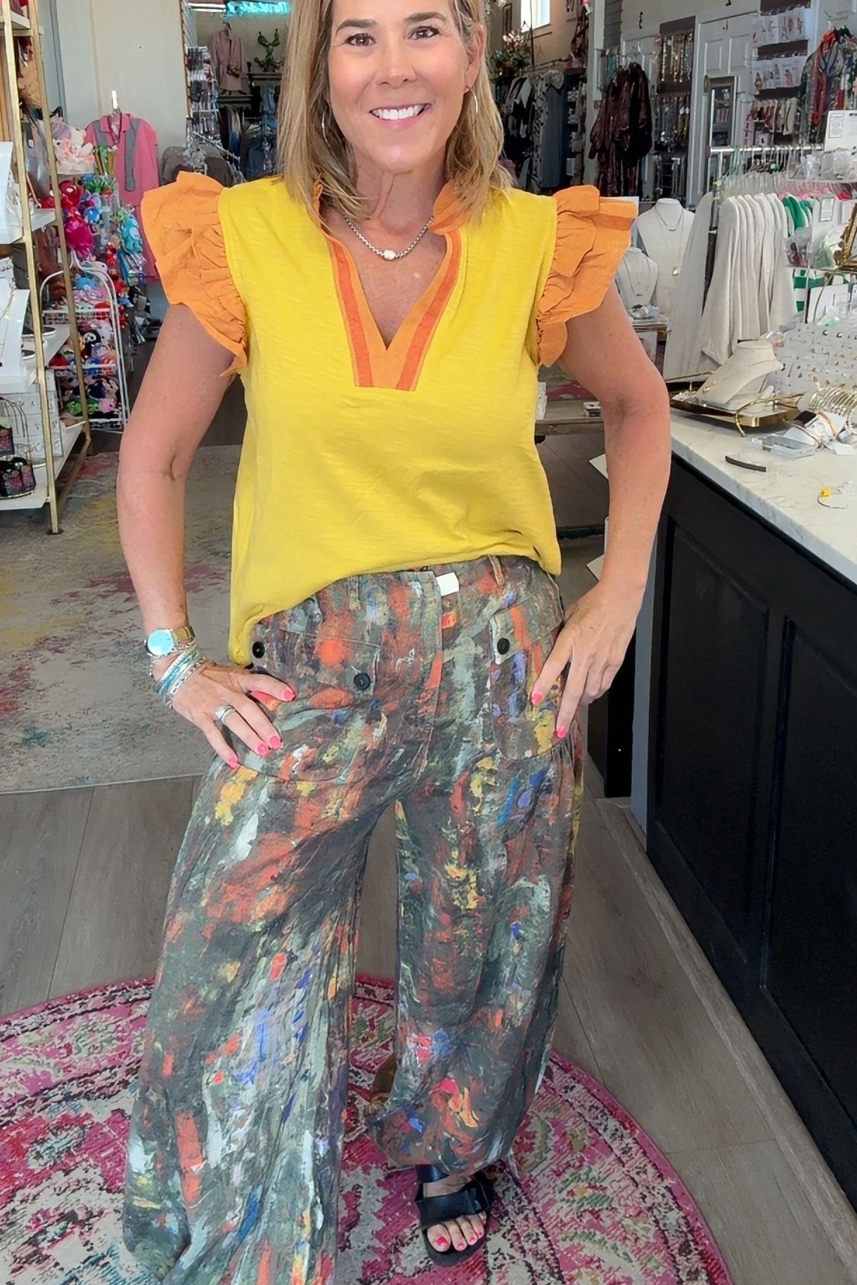 A woman posing inside a boutique shop, wearing a bright yellow ruffle-sleeve top with orange trim and wide-leg, multi-colored abstract print pants.