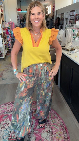 A woman posing inside a boutique shop, wearing a bright yellow ruffle-sleeve top with orange trim and wide-leg, multi-colored abstract print pants.