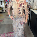 A woman stands inside a boutique shop wearing a long beige fringed dress with a floral print. The dress features a light beige collar and a long, layered fringe skirt with shades of brown, red, and blue.