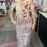 A woman stands inside a boutique shop wearing a long beige fringed dress with a floral print. The dress features a light beige collar and a long, layered fringe skirt with shades of brown, red, and blue.