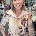 A woman with blonde hair stands in a retail store wearing a beige fringed blouse with a vintage-style floral print featuring red roses and black leaves.