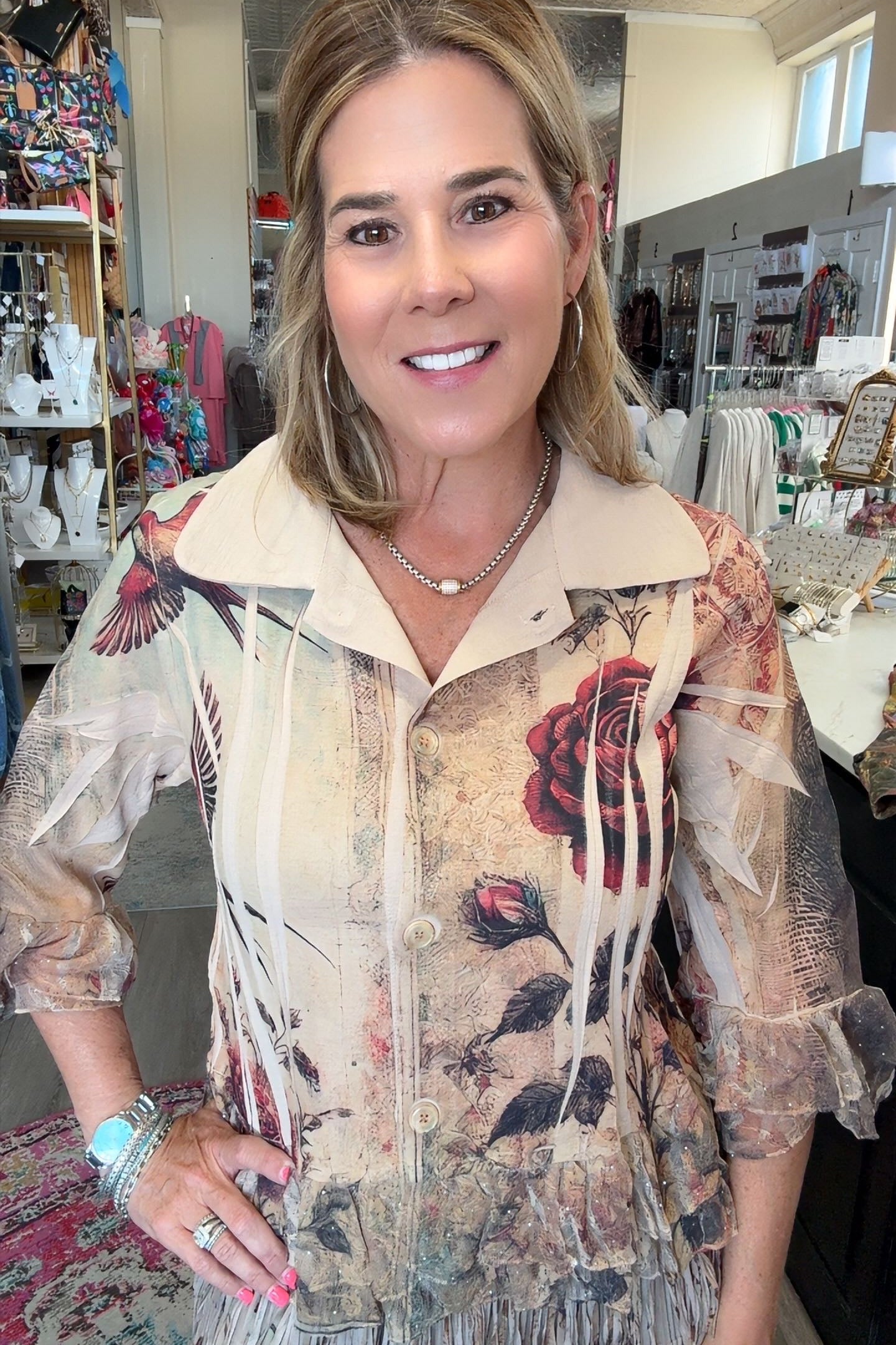 A woman with blonde hair stands in a retail store wearing a beige fringed blouse with a vintage-style floral print featuring red roses and black leaves.