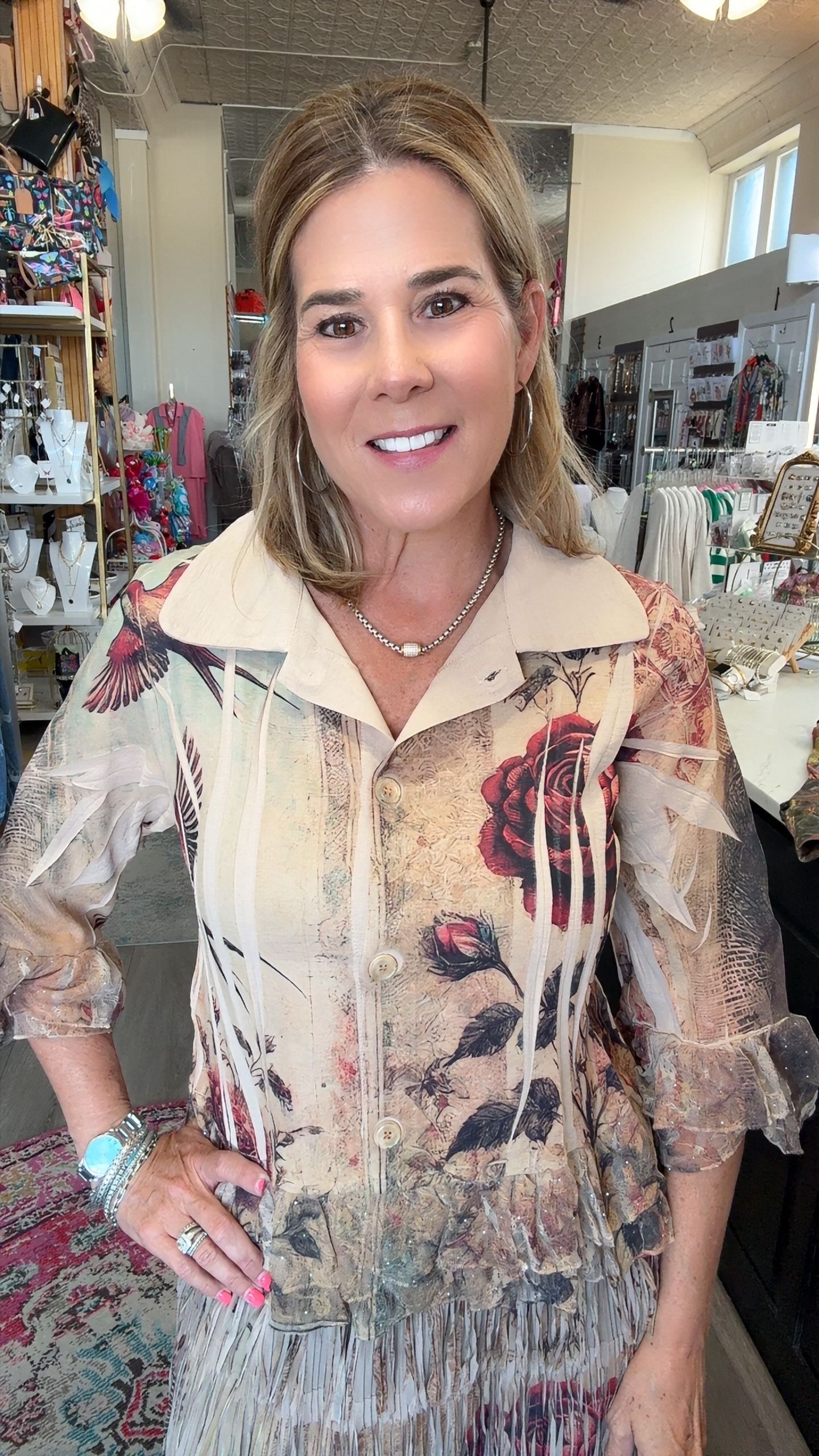 A woman with blonde hair stands in a retail store wearing a beige fringed blouse with a vintage-style floral print featuring red roses and black leaves.
