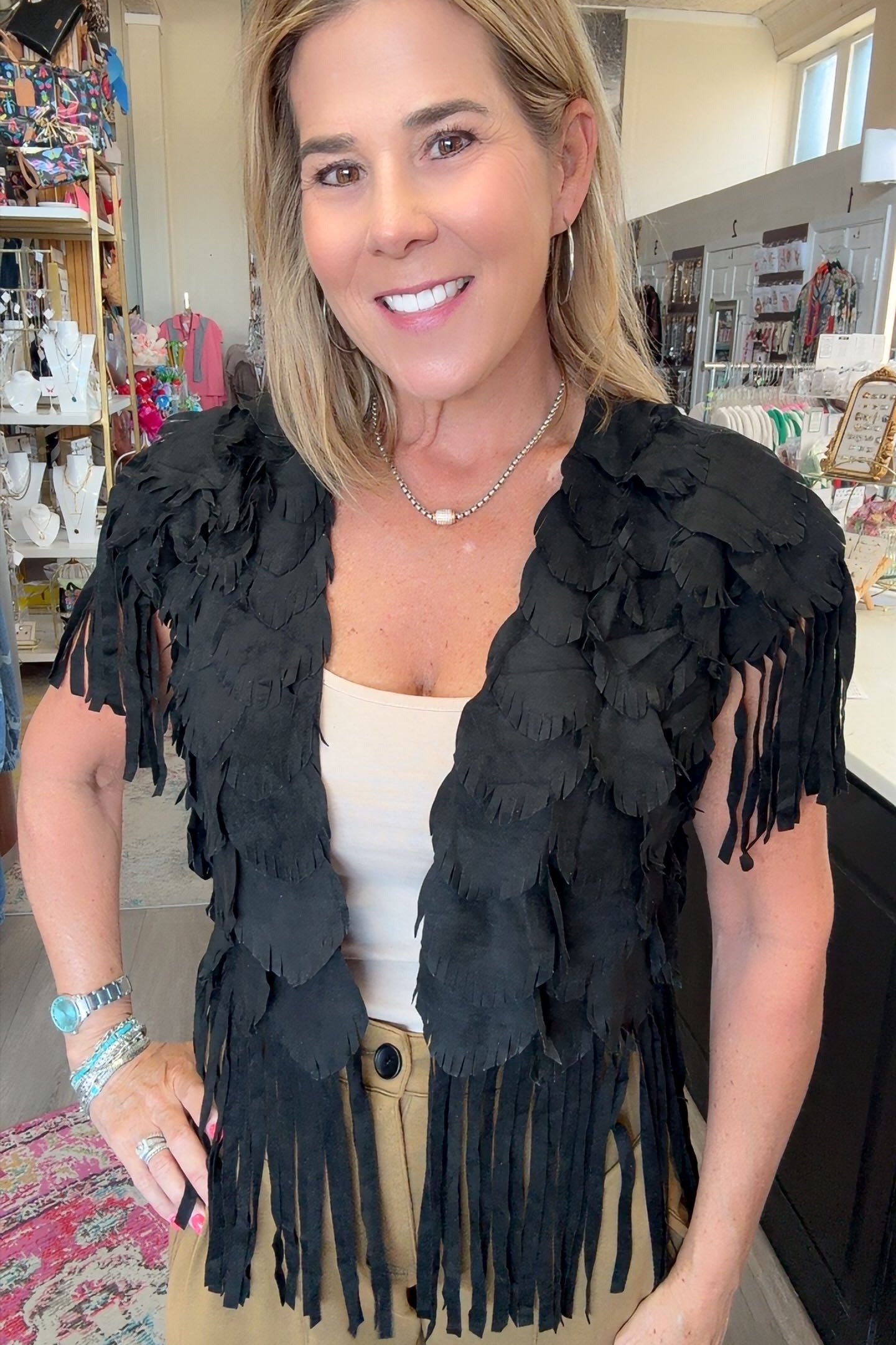 A woman wearing a black feathered fringe vest over a cream top and khaki pants, standing in a retail store interior.