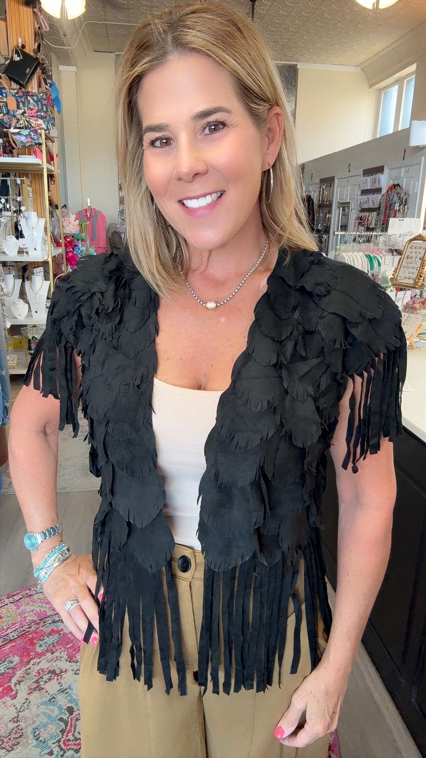 A woman wearing a black feathered fringe vest over a cream top and khaki pants, standing in a retail store interior.