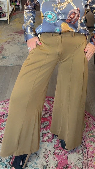 A woman posing in a boutique shop, wearing a blue patterned long-sleeve button-up shirt and wide-leg tan pants.