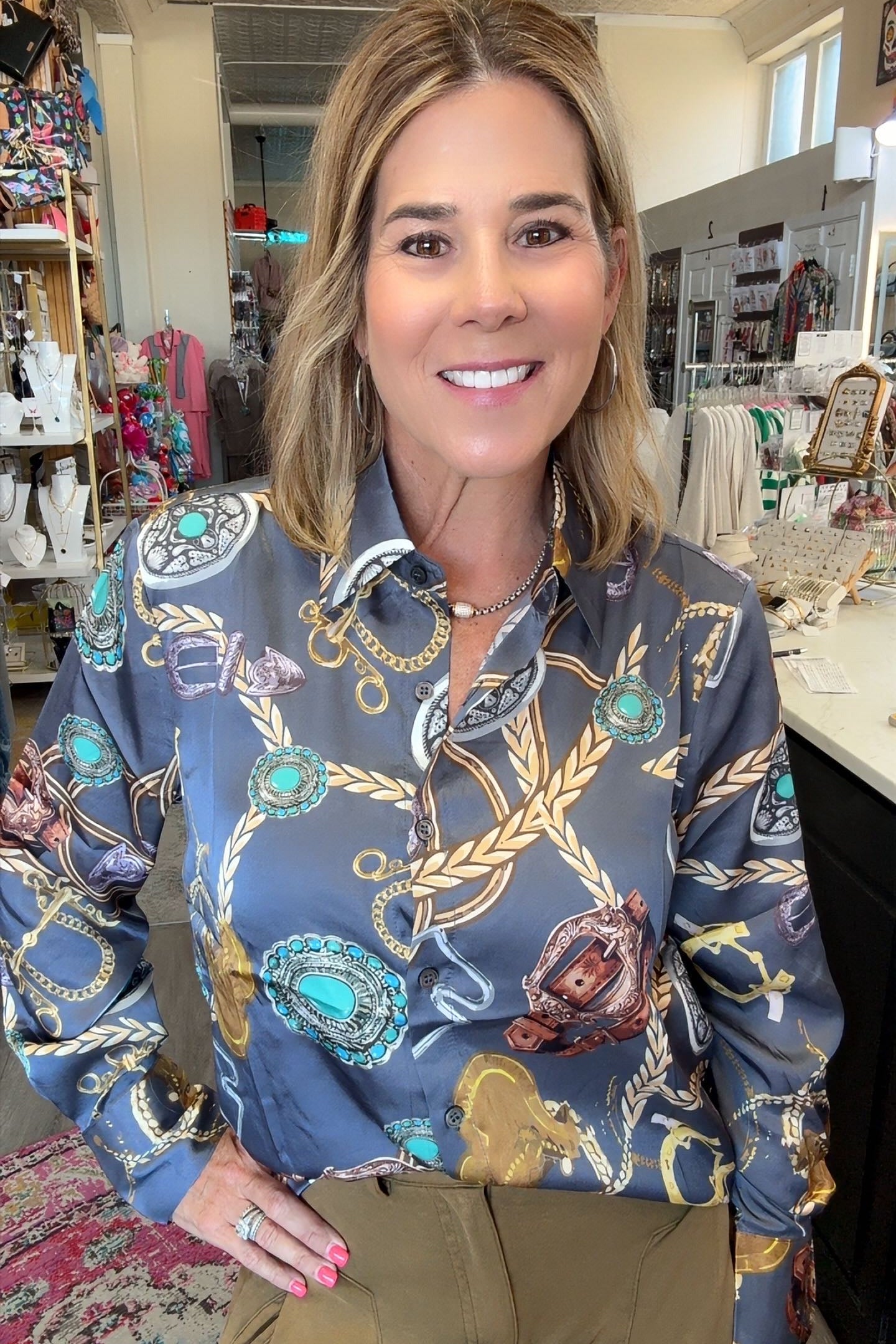 A woman smiling in a boutique while wearing a long-sleeve button-up shirt with a detailed gold, turquoise, and brown rope and chain pattern on a slate blue background, paired with olive green pants.