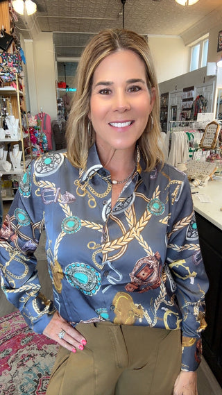 A woman smiling in a boutique while wearing a long-sleeve button-up shirt with a detailed gold, turquoise, and brown rope and chain pattern on a slate blue background, paired with olive green pants.