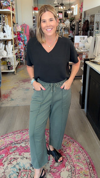 A woman stands in a boutique shop wearing a black short-sleeve v-neck top and sage green drawstring pants. The shop interior features light wood floors, a pink patterned rug, and shelves and counters filled with various clothing items.