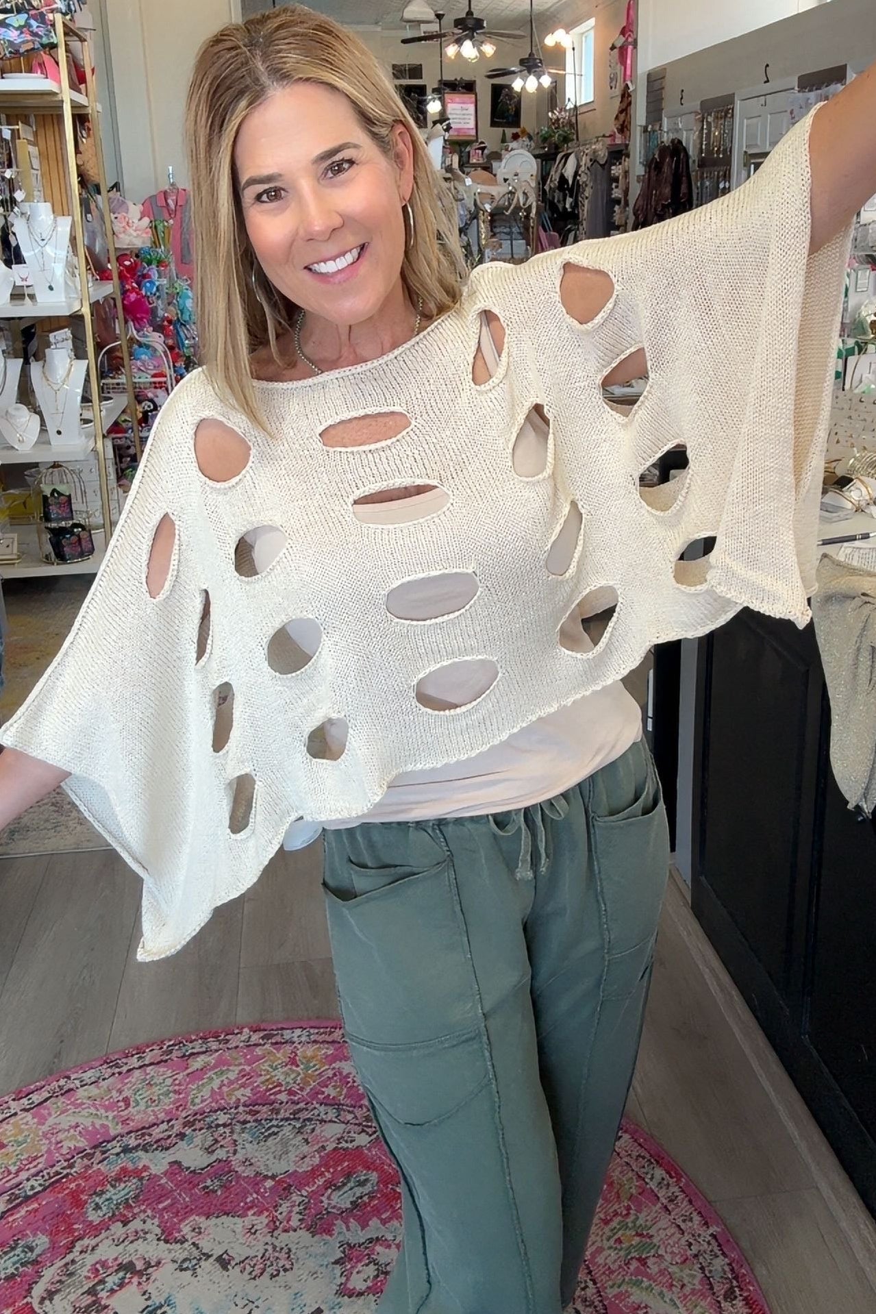 A woman modeling a cream-colored, open-knit poncho-style top with large cutout details, paired with sage green pants, standing inside a boutique shop.