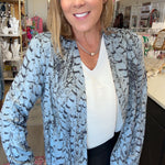 A woman posing inside a boutique shop, wearing a light blue and gray abstract patterned blazer over a white ribbed top and black pants.