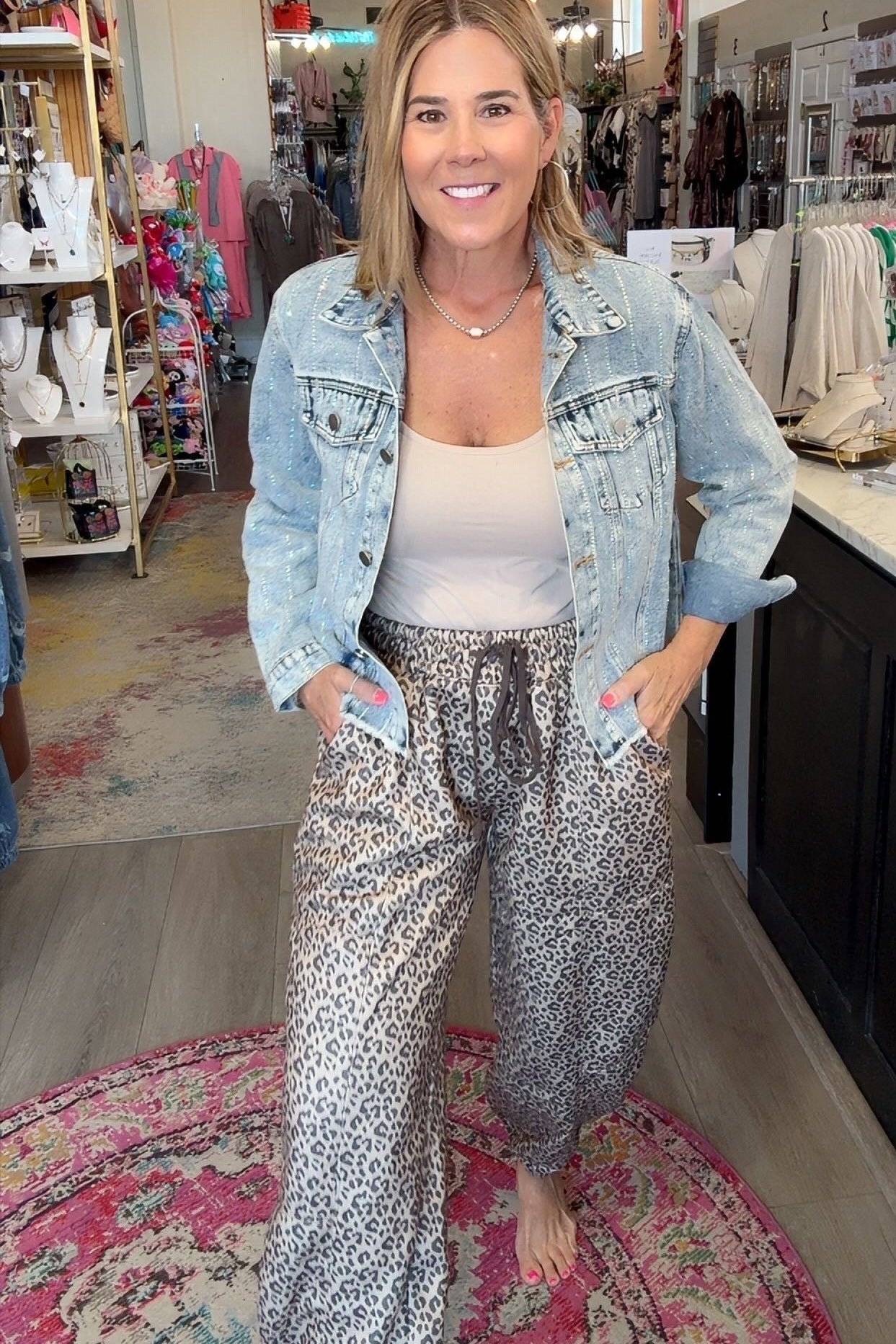 A woman stands barefoot in a boutique shop wearing a light-wash denim jacket, a white tank top, and loose gray leopard-print pants. She is positioned on a pink patterned rug with store fixtures, clothing racks, and displays visible in the background.