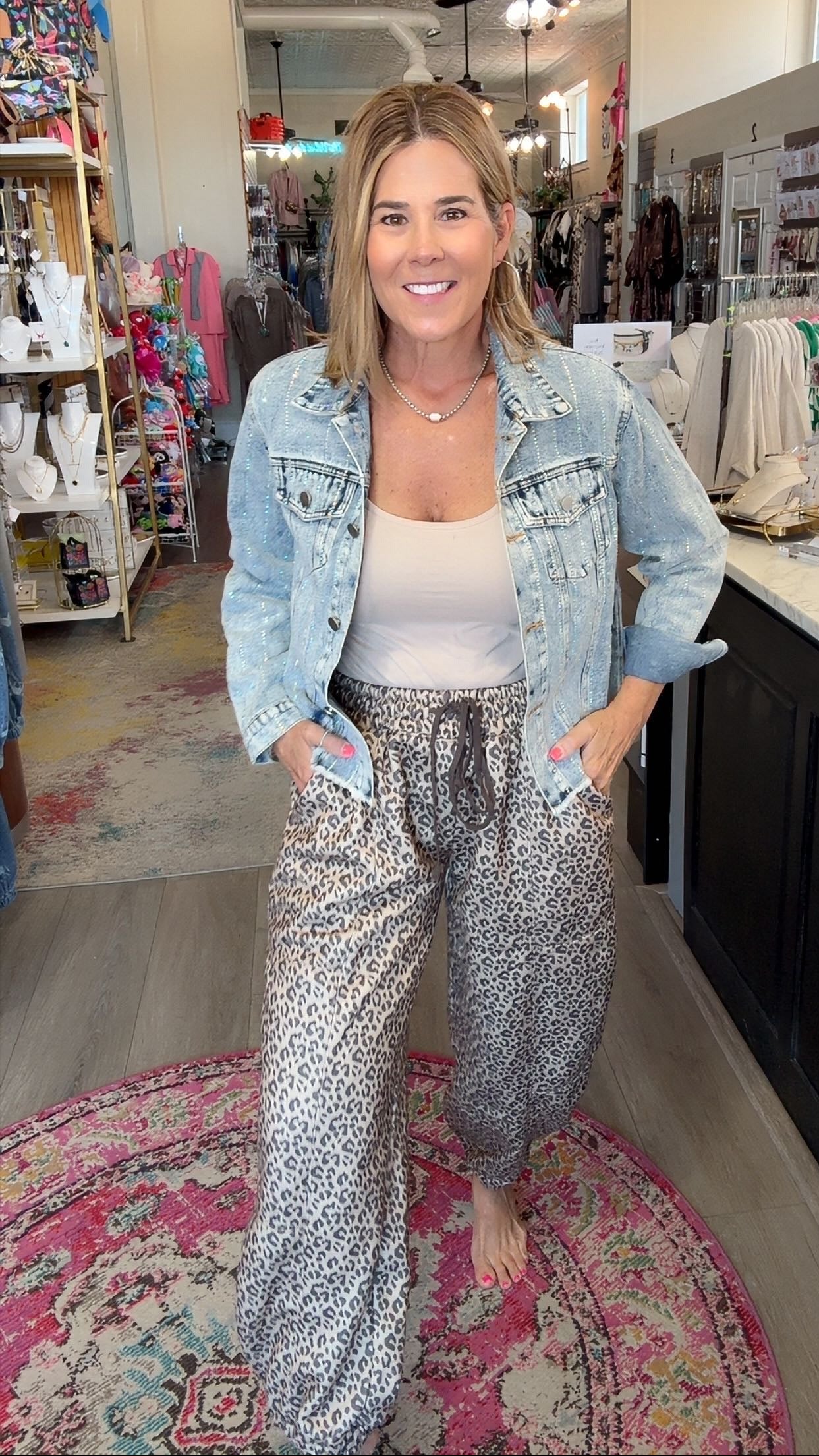 A woman stands barefoot in a boutique shop wearing a light-wash denim jacket, a white tank top, and loose gray leopard-print pants. She is positioned on a pink patterned rug with store fixtures, clothing racks, and displays visible in the background.