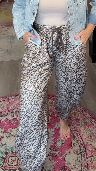 A woman stands barefoot in a boutique shop wearing a light-wash denim jacket, a white tank top, and loose gray leopard-print pants. She is positioned on a pink patterned rug with store fixtures, clothing racks, and displays visible in the background.