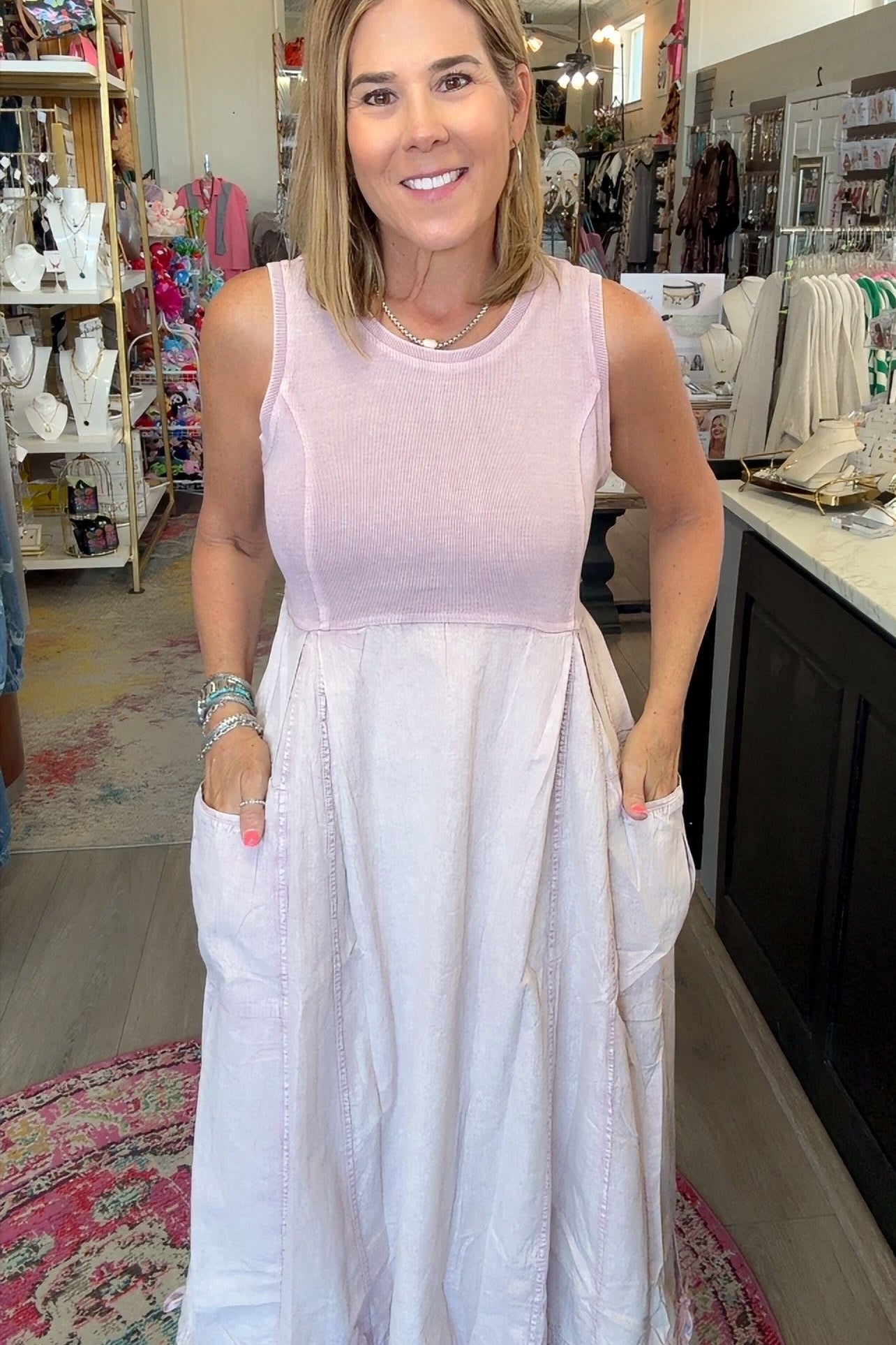 A woman wearing a sleeveless two-tone dress with a light pink bodice and a white skirt with side pockets, standing inside a retail boutique.