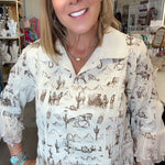 A smiling woman with blonde hair wearing a cream-colored long-sleeve blouse with a brown western desert print, paired with blue jeans, standing inside a retail store.