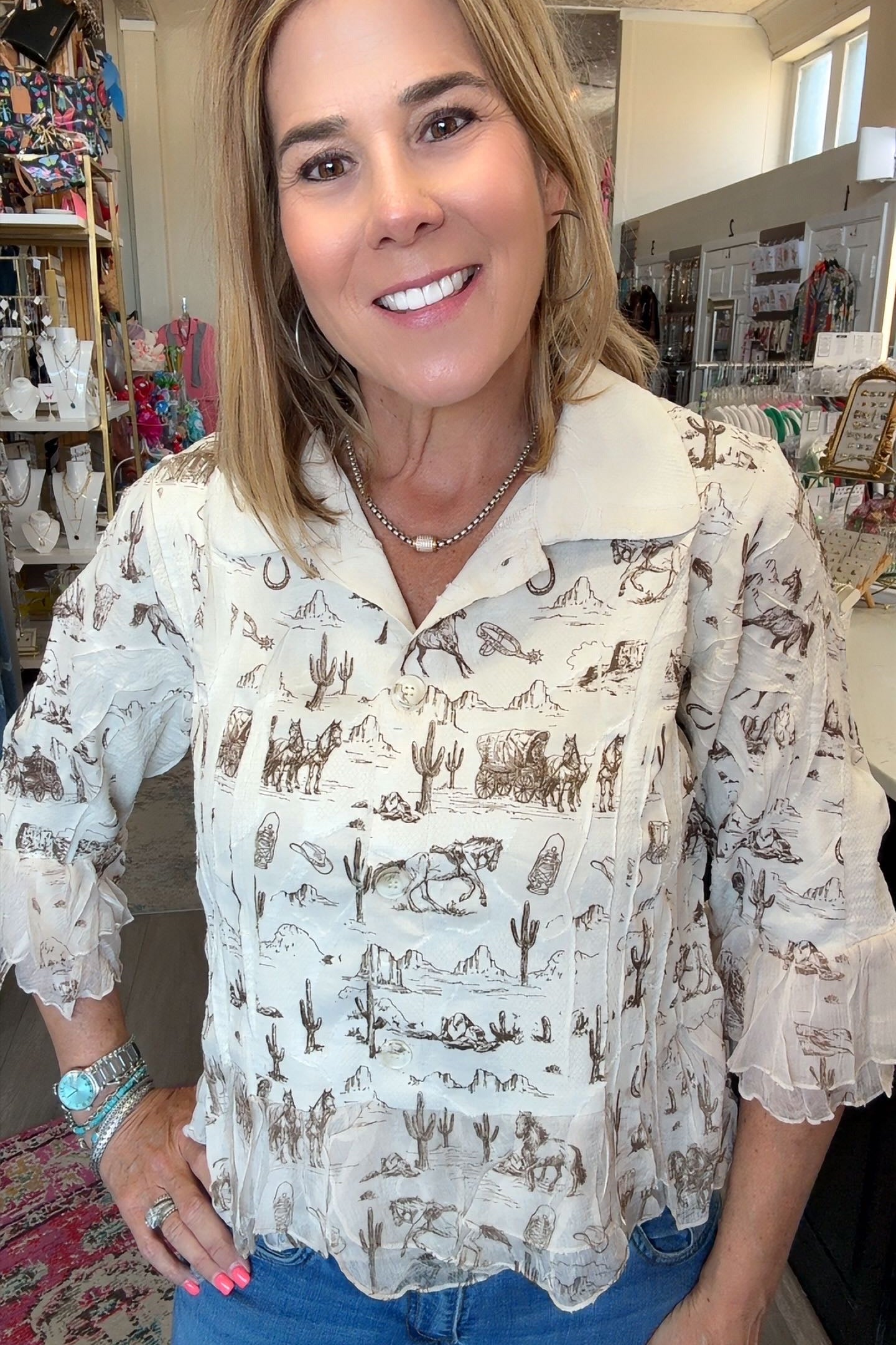 A smiling woman with blonde hair wearing a cream-colored long-sleeve blouse with a brown western desert print, paired with blue jeans, standing inside a retail store.