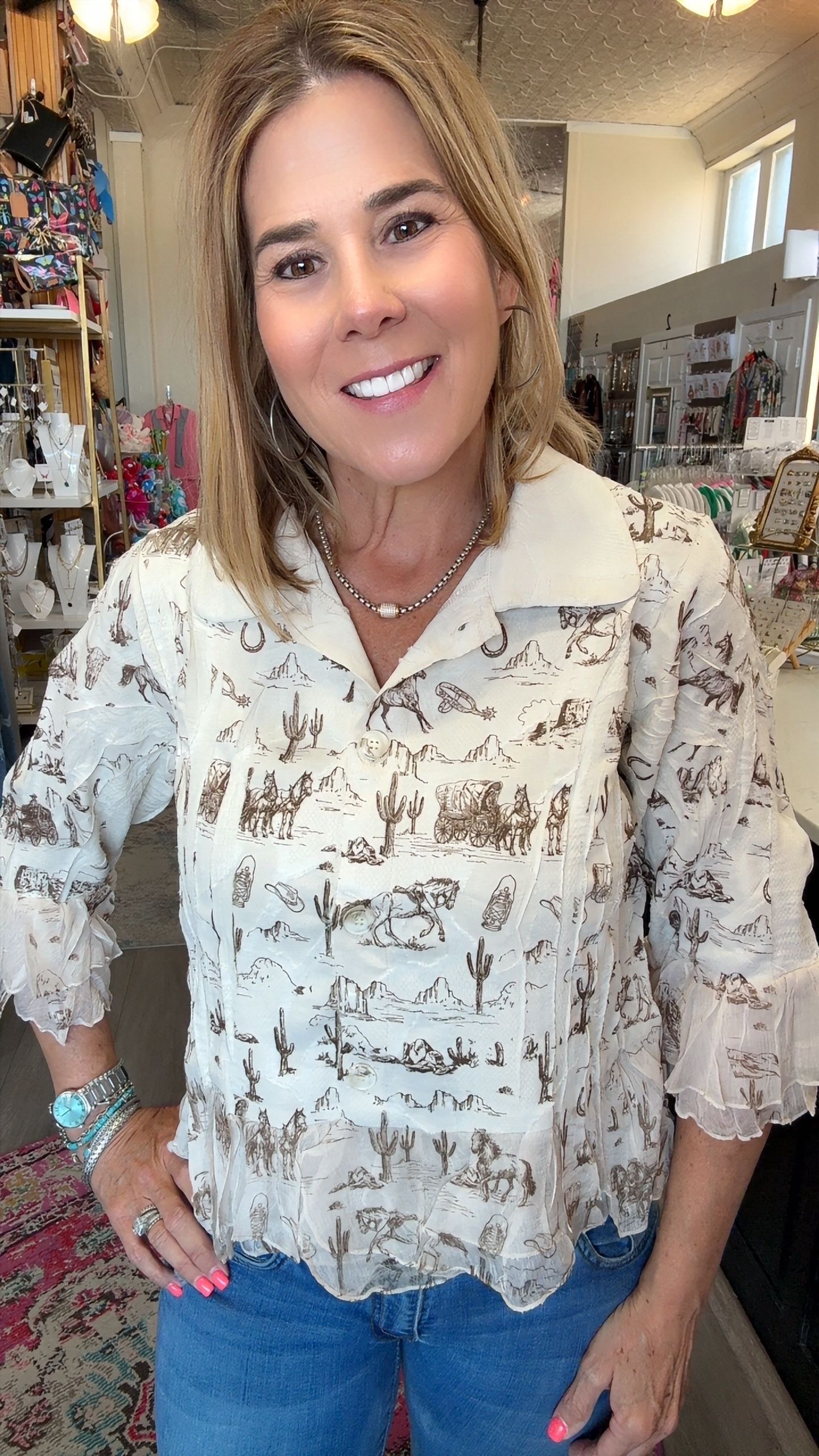 A smiling woman with blonde hair wearing a cream-colored long-sleeve blouse with a brown western desert print, paired with blue jeans, standing inside a retail store.