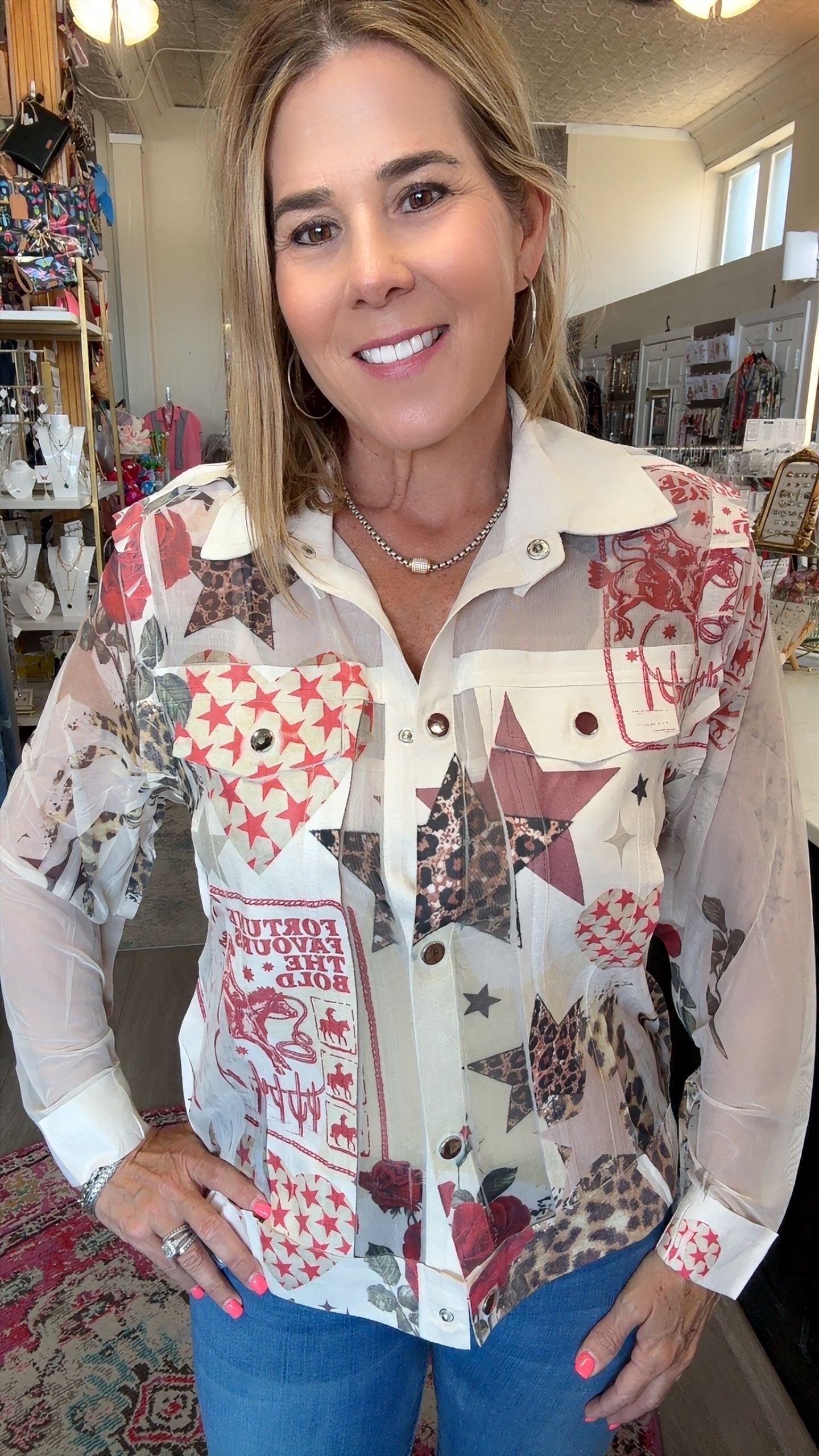 A smiling woman wearing a long-sleeve button-up shirt with a patchwork print featuring red, beige, brown, and white patterns, paired with blue jeans in what appears to be a retail store.