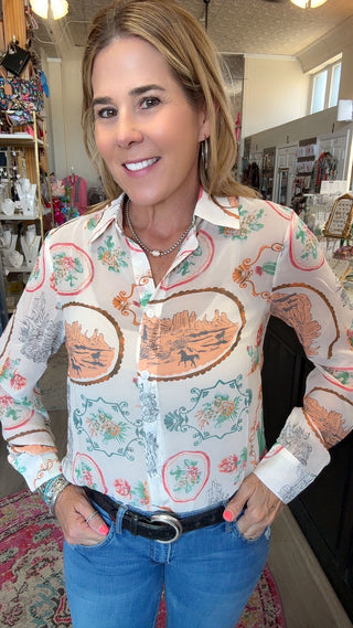 A woman wearing a long-sleeve button-up blouse with a vintage-style landscape and floral pattern, paired with blue jeans and a black belt, standing inside a retail store.