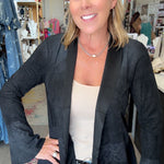 A woman posing in a clothing boutique wearing a black textured open-front blazer over a beige tank top and blue jeans.