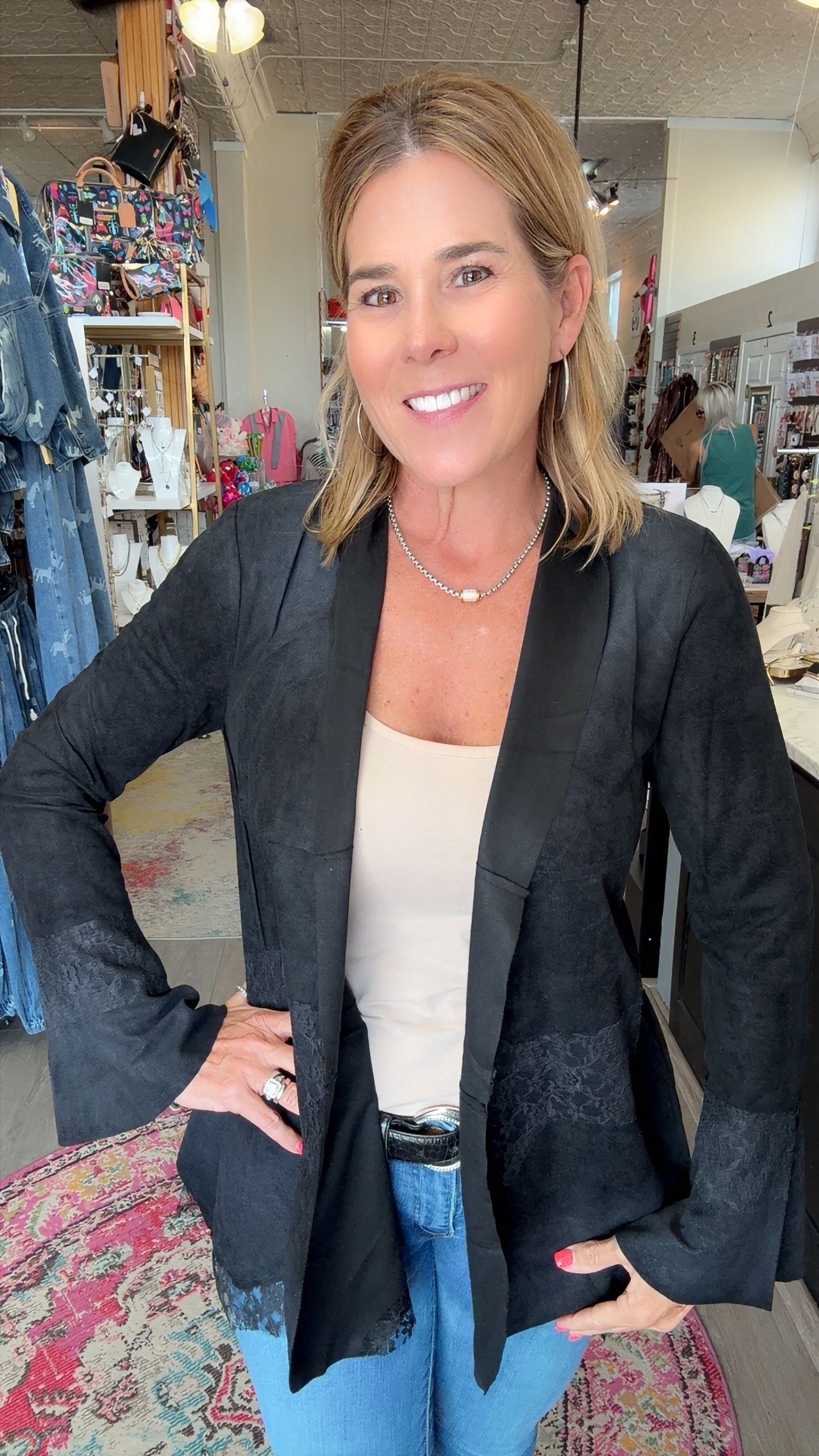 A woman posing in a clothing boutique wearing a black textured open-front blazer over a beige tank top and blue jeans.
