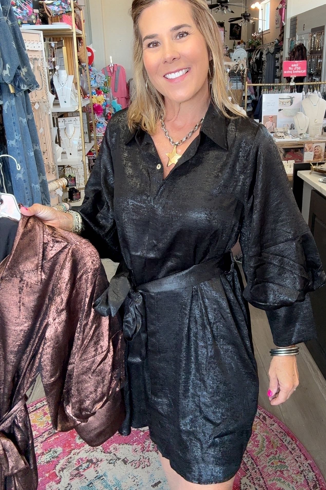 A woman stands inside a clothing boutique wearing a black textured mini shirt dress with puff sleeves and a tie waist. She is holding up a brown satin robe on a hanger to the left.