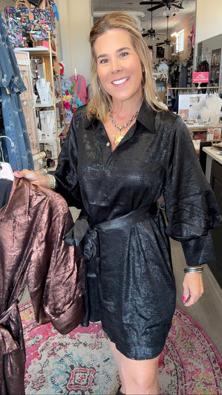 A woman stands inside a clothing boutique wearing a black textured mini shirt dress with puff sleeves and a tie waist. She is holding up a brown satin robe on a hanger to the left.