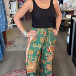 A woman posing in a boutique shop, wearing a black tank top and green wide-leg pants with a colorful floral pattern and striped hem.