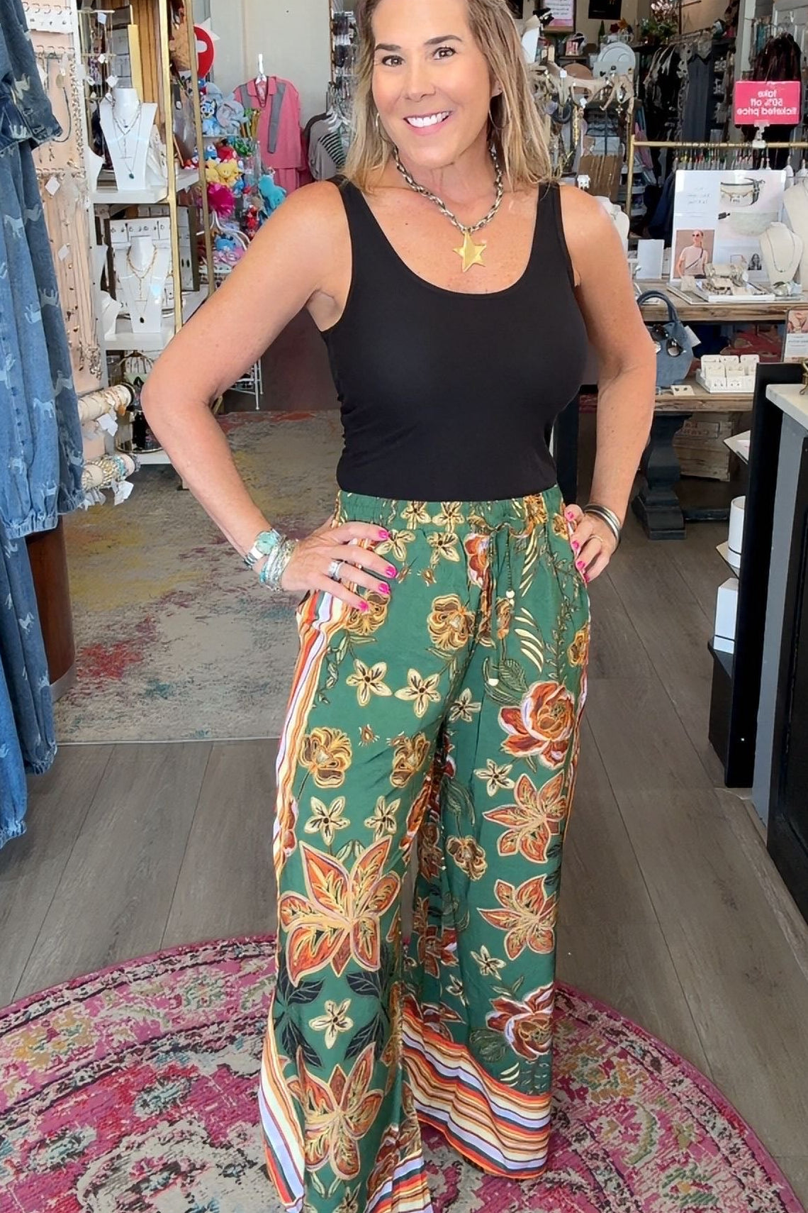 A woman posing in a boutique shop, wearing a black tank top and green wide-leg pants with a colorful floral pattern and striped hem.