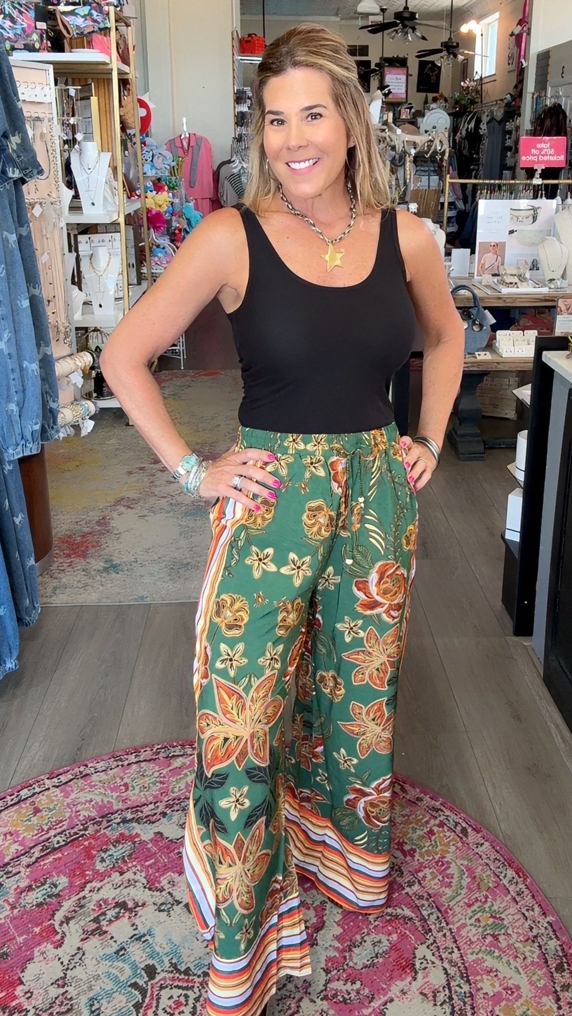 A woman posing in a boutique shop, wearing a black tank top and green wide-leg pants with a colorful floral pattern and striped hem.