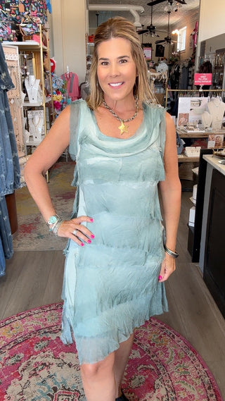 A woman wearing a sleeveless, tiered light blue-green fringe dress stands inside a boutique shop, posing on a patterned rug.