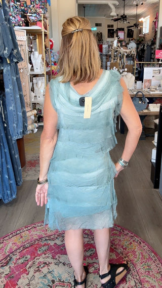 A woman viewed from behind wearing a light blue tiered tulle dress inside a clothing boutique, standing on a patterned rug.