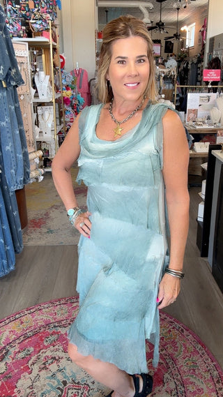 A woman wearing a light blue sleeveless draped dress stands inside a retail boutique, posing on a patterned rug with store fixtures visible in the background.