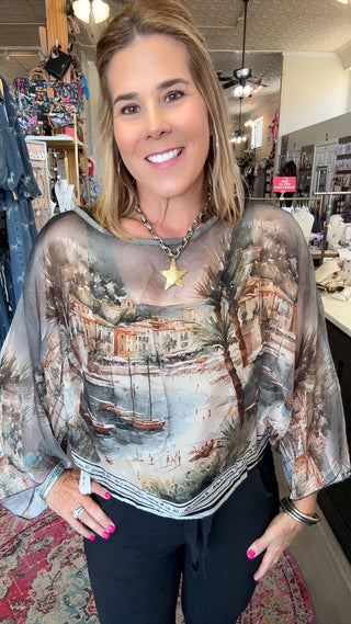 A woman with blonde hair smiling in a boutique, wearing a sheer, semi-sheer long-sleeve top with a scenic coastal town print in muted earth tones of brown, beige, and gray, paired with black pants.