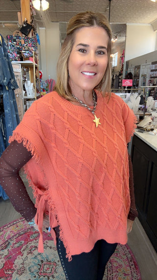 A woman modeling a coral orange knit poncho sweater with fringe and a diamond cable pattern, layered over sheer dark brown dotted long-sleeve sleeves, in a retail store setting.
